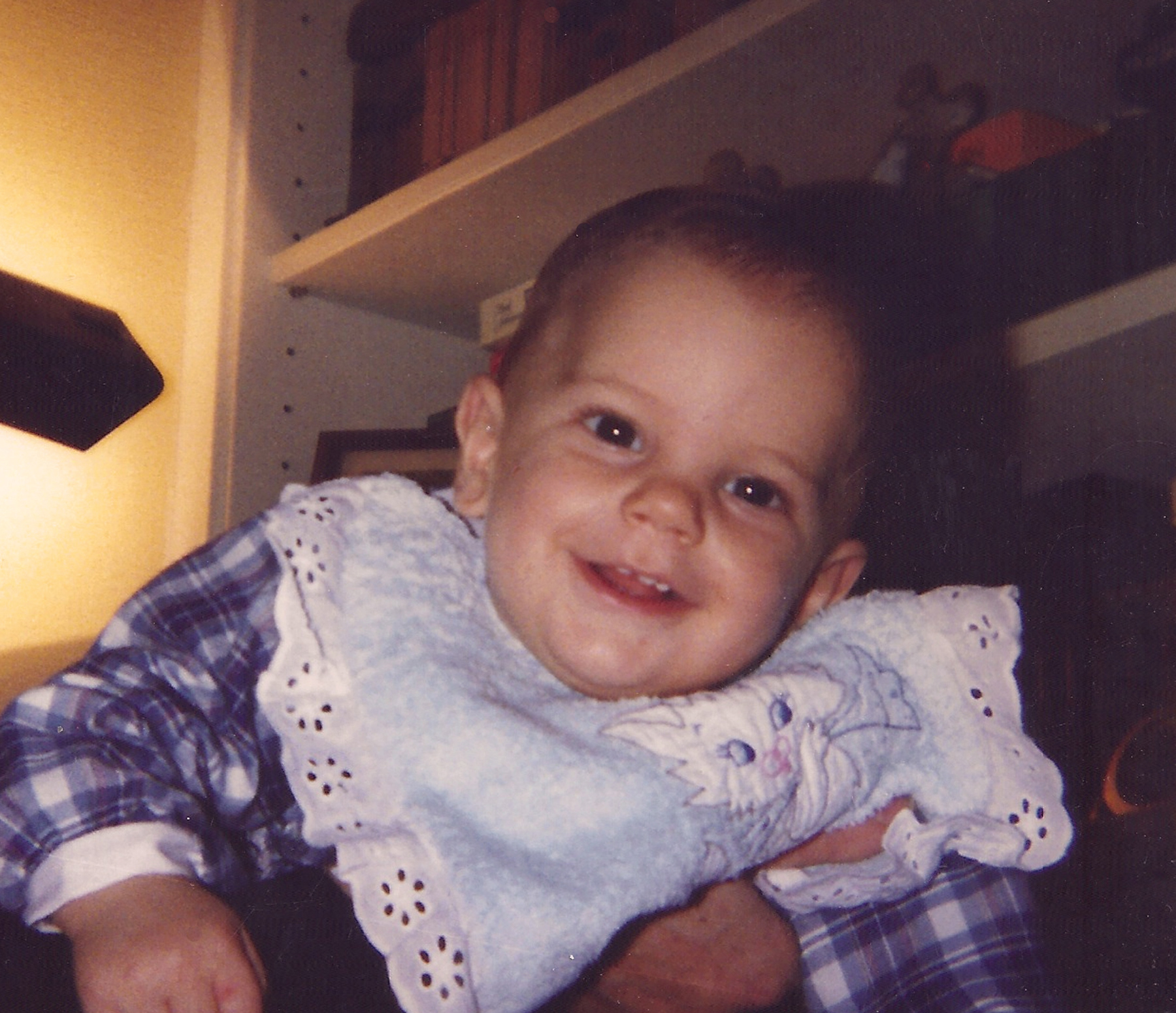 Carlo Acutis als Baby in einem blauen karierten Body, mit einem fröhlichen Blick. Das Bild zeigt ihn in den frühen Lebensjahren.